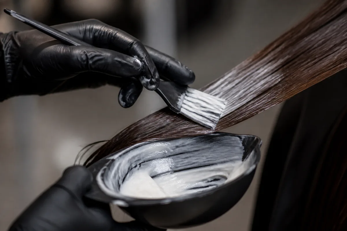 Mains gantées d’une coiffeuse appliquant un soin lissant mèche par mèche sur une chevelure brune, cadrage serré sur le peigne