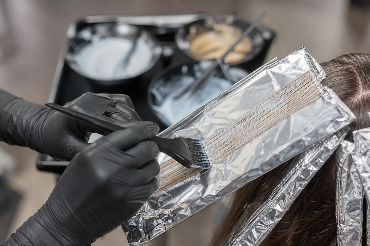 Mains gantées d&rsquo;une coloriste appliquant un balayage à l&rsquo;aide de bandes de papier aluminium, bols de coloration posés à côté sur une tablette de salon